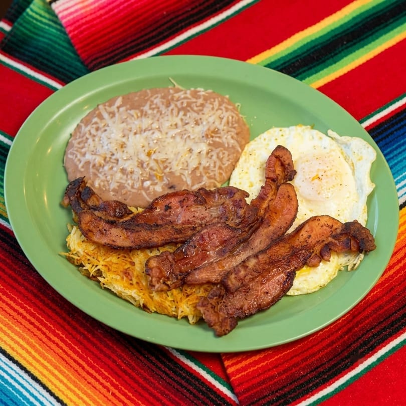 Breakfast plate with bacon, fried eggs, hash browns, and refried beans on a green plate.