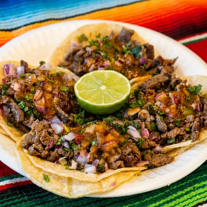 Three street tacos with carne asada, red onion, cilantro, salsa, and a lime wedge.