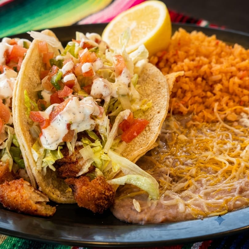 Two fish tacos with lettuce, pico de gallo, and crema, alongside rice and refried beans.